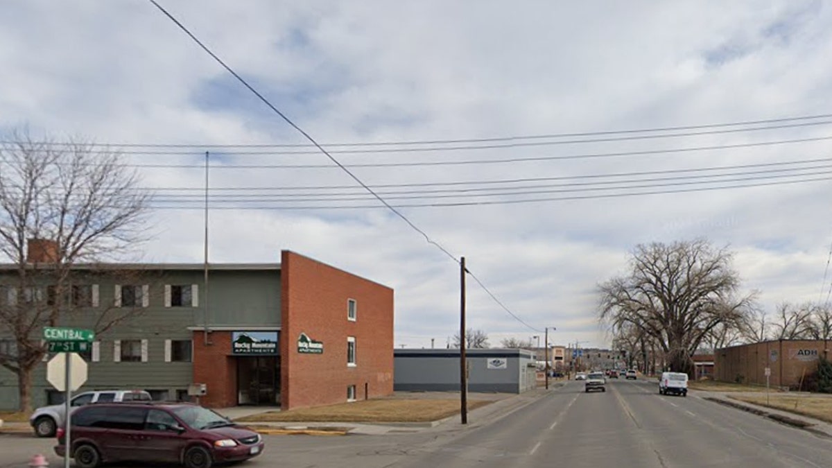 A minivan stopped at a stop sign next to a Rocky Mountains apratment building at an intersection