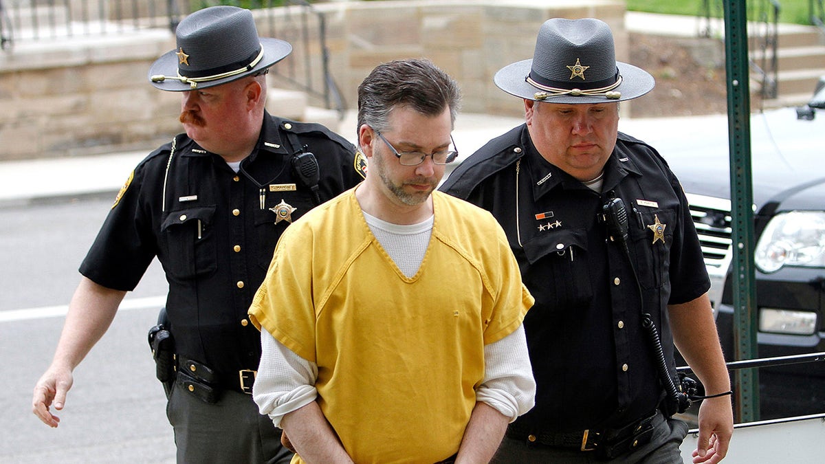 Shawn Grate in a yellow and white jumpsuit being escorted by two policemen.