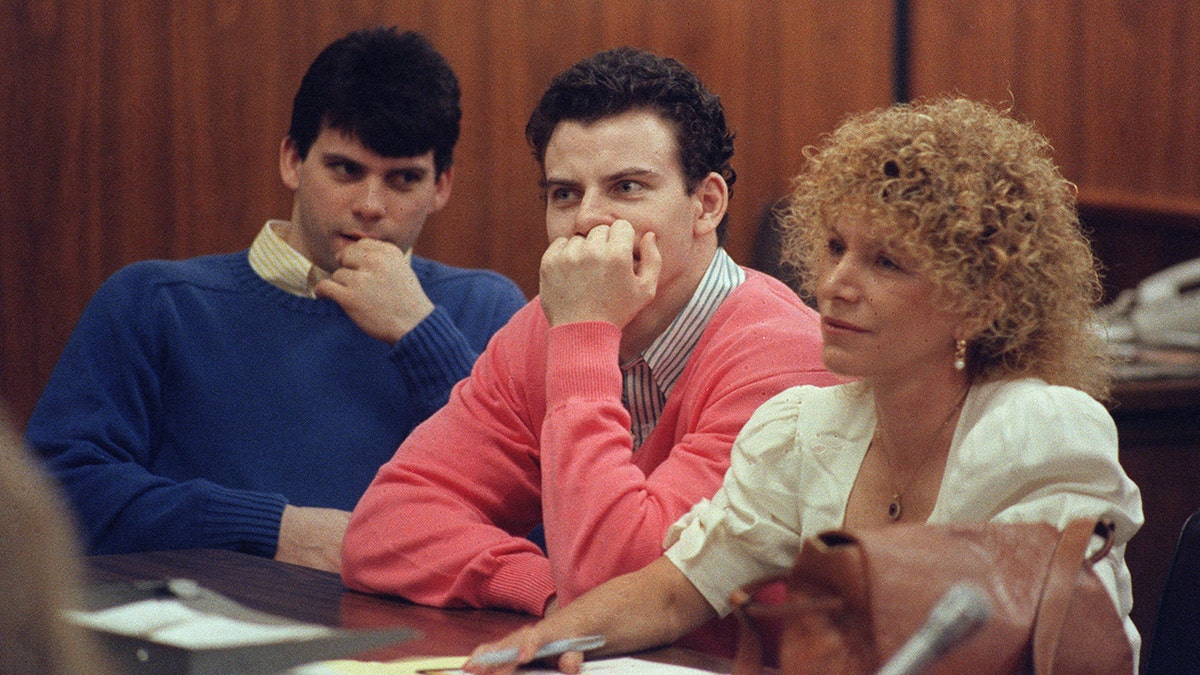 Lyle Menendez in a blue sweater and Kyle Menendez in a coral sweater sit with attorney Leslie Abramson, both putting hands to their mouths and chin