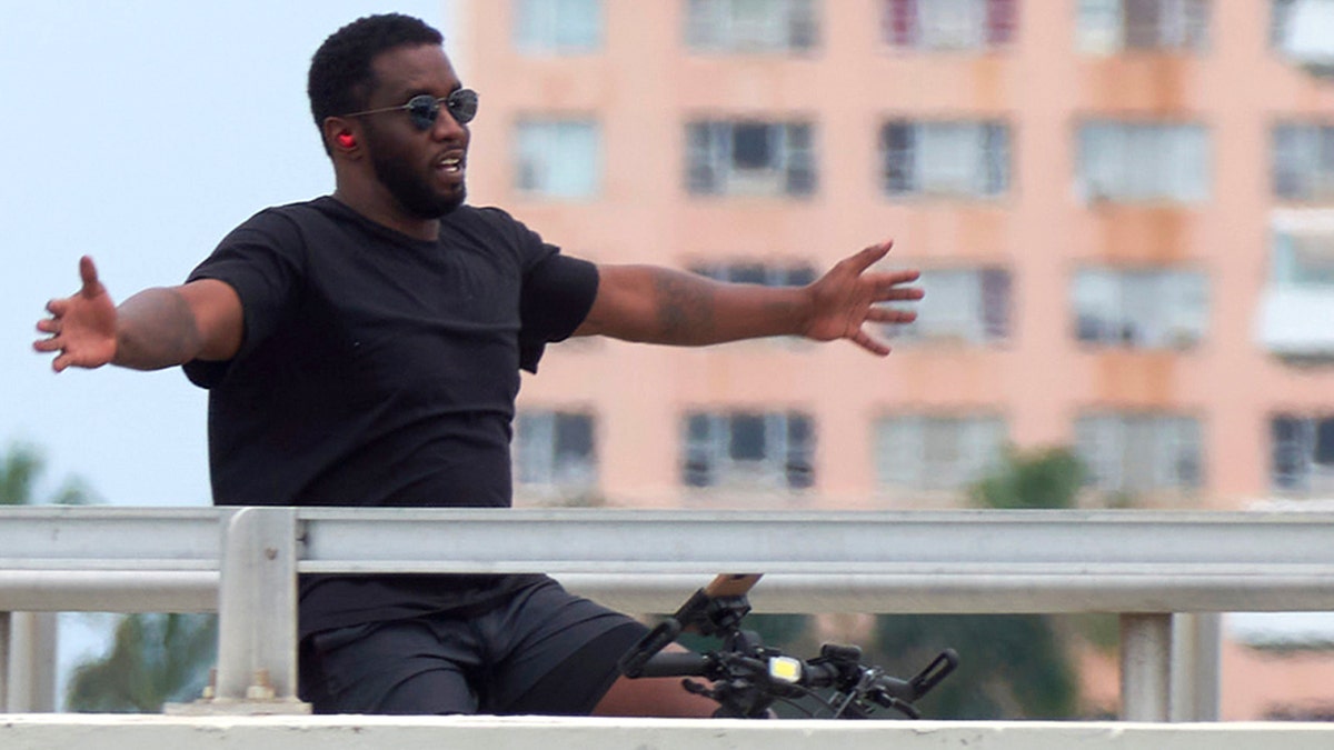 P Diddy rides has bicycle with arms outstretched wearing sunglasses.