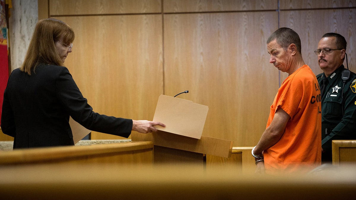 Jeremy Scott in court wearing an orange jumpsuit looking at a file folder.
