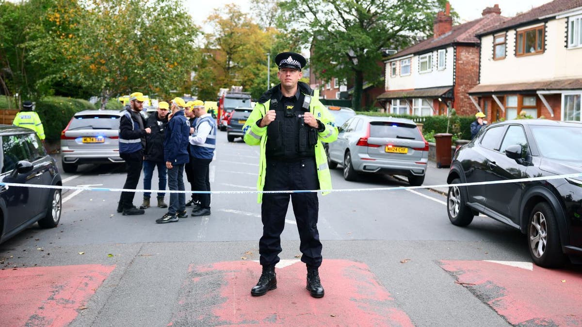 Police officer in high-visibility jacket stands behind cordon near houses after Manchester synagogue attack.