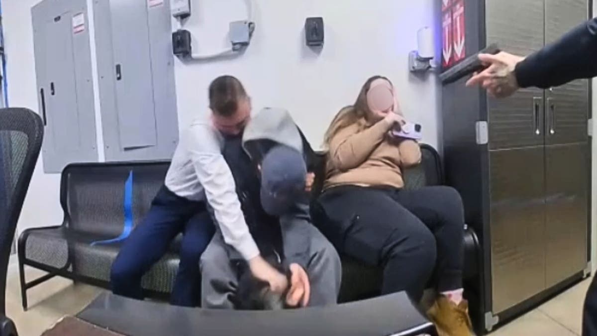 A security worker and police officer restrain a suspect on a bench inside a store security room as a woman recoils nearby.