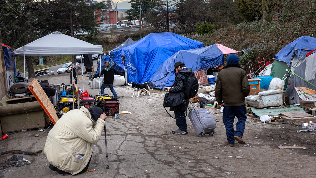 People walk through a homeless encampment with tents, tarps, and belongings along a paved roadway